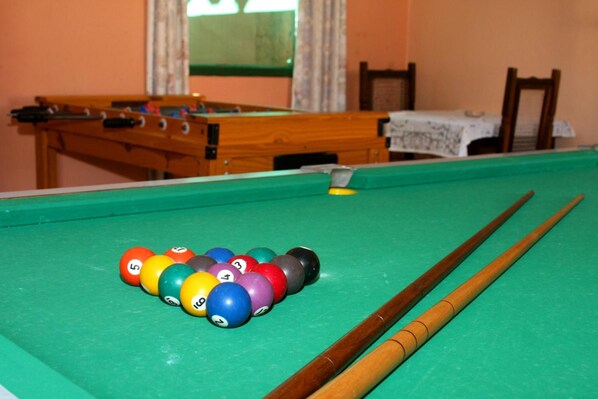 A pool table with billiard balls and cues, a wooden table tennis table, and a chair in a room with a window and a tablecloth. - Pousada Marinamarela (Penha)
