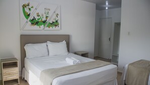 A neatly made double bed with white linens, a wooden nightstand, a small bedside table, and a wall-mounted abstract art piece. - Pousada Marinamarela (Penha)