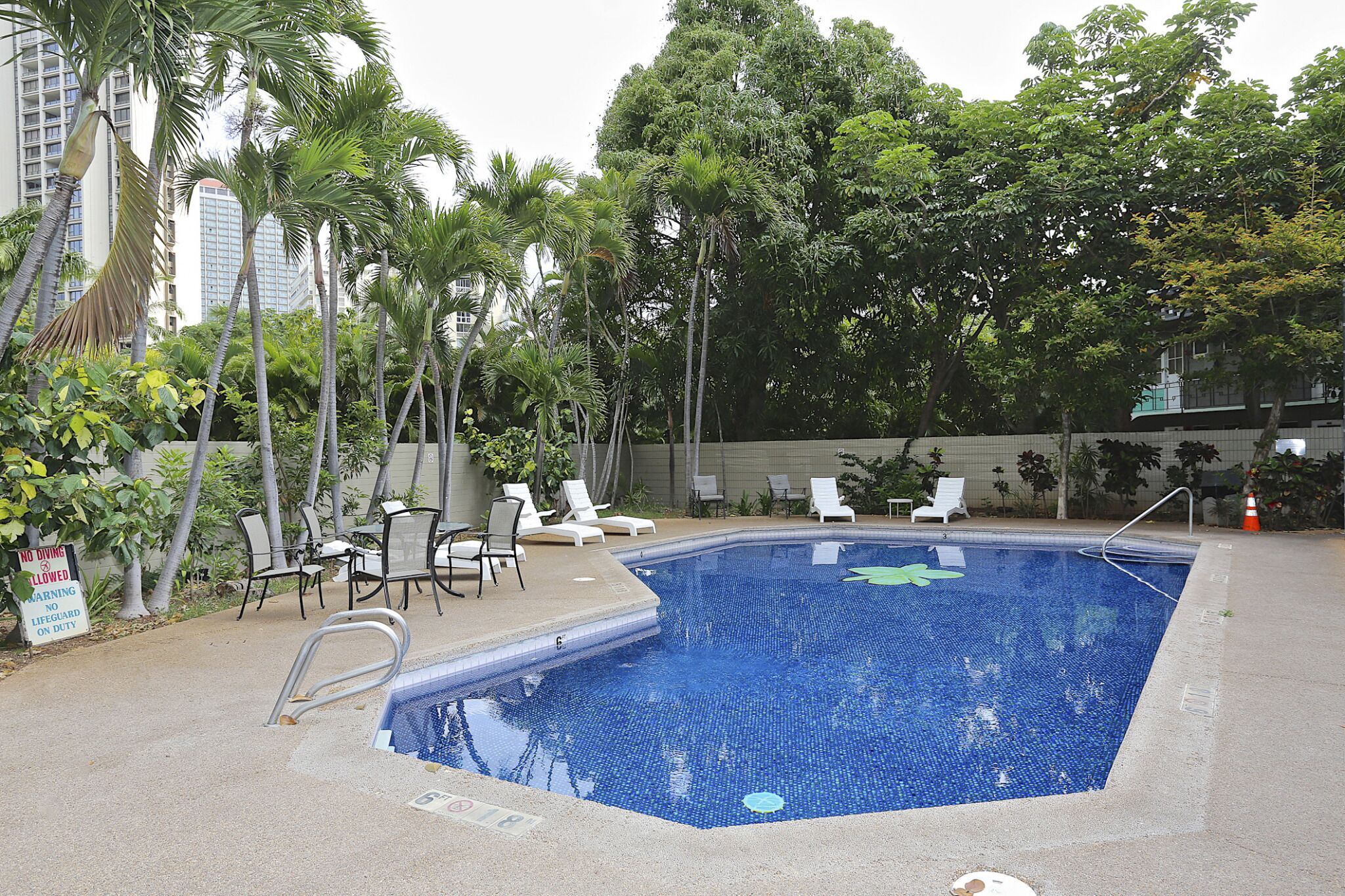 Une piscine entourée de palmiers, avec des chaises longues et un poste de surveillance pour le sauveteur, et un bâtiment visible au loin.
