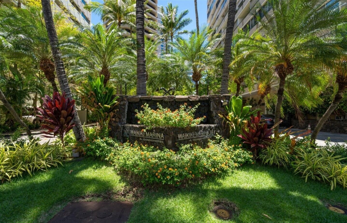 A lush garden with a stone sign for Waikiki Marina A Condominium Hotel surrounded by tropical plants and palm trees.