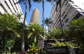 Un immeuble imposant à l’architecture moderne, entouré de palmiers et d’un mur de pierre sur lequel on peut lire l’inscription « Waikiki Marina Condo Hotel ».