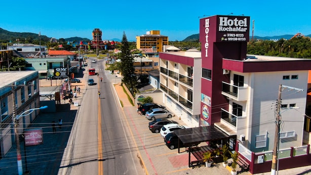 A hotel with a prominent sign, a parking lot, and a street view with buildings and mountains in the background. - Hotel Rochamar (Penha)