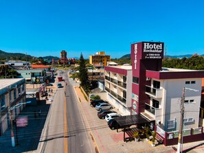 A hotel with a prominent sign, a parking lot, and a street view with buildings and mountains in the background. - Hotel Rochamar (Penha)