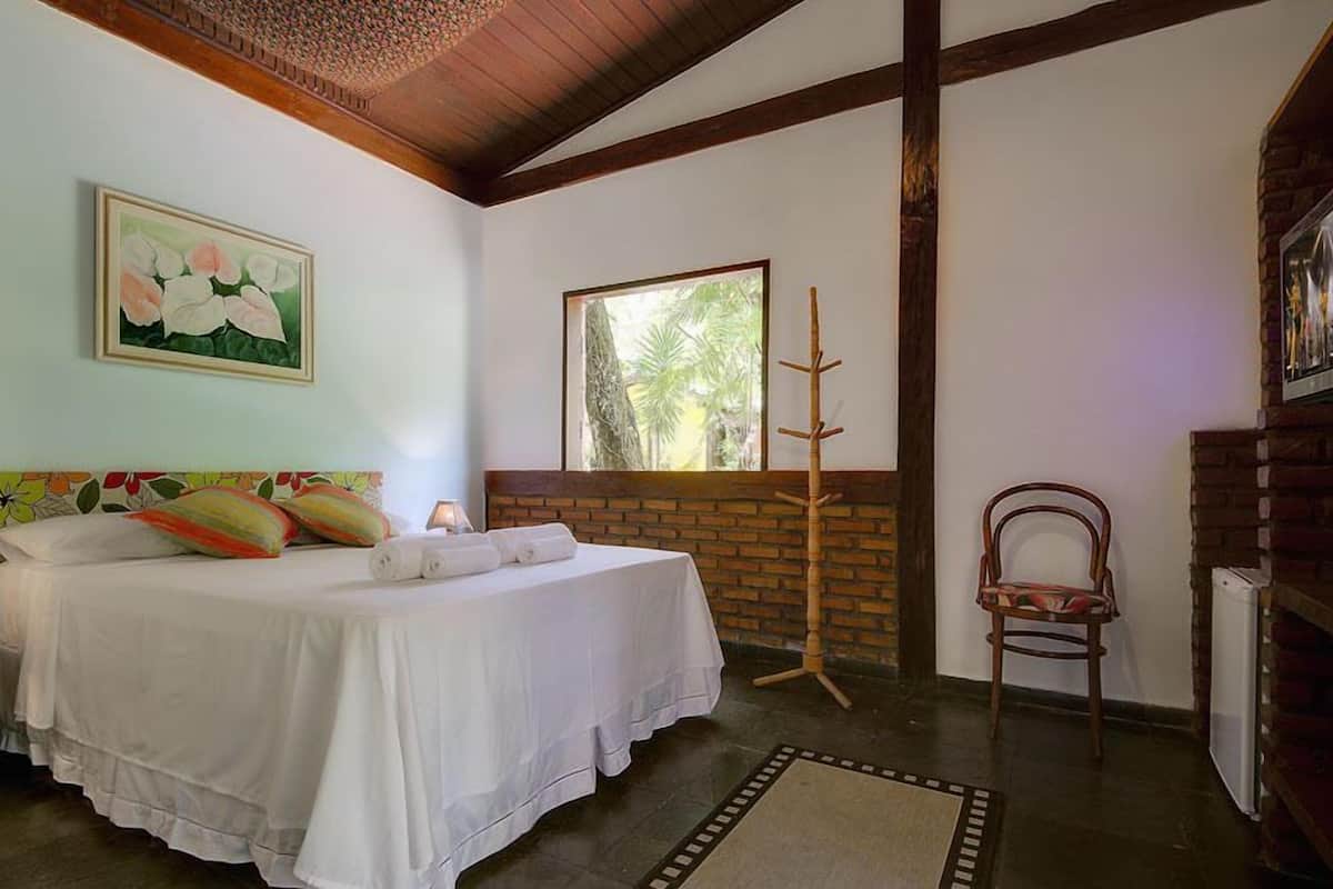 A bedroom with a bed, a window with a view of greenery, a wooden chair, and a brick wall.