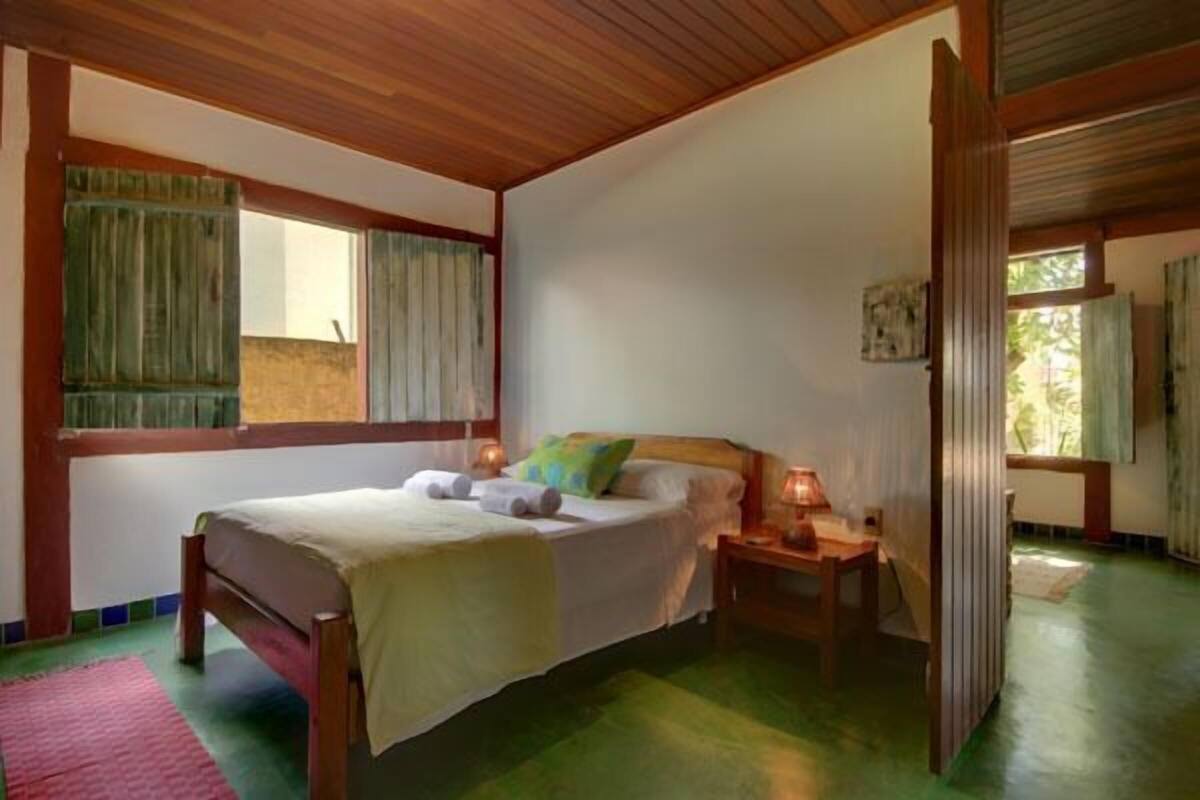 A bedroom with a wooden bed, a nightstand with a lamp, a window with curtains, and a green floor.