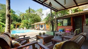 Área da piscina com mobiliário de madeira, um espaço de estar coberto e vista para a piscina e vegetação ao redor.