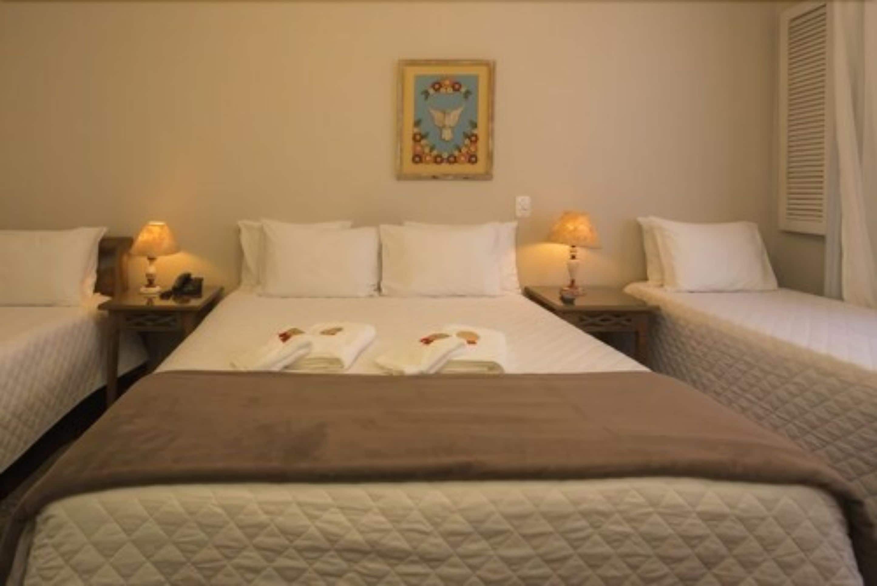 Une chambre d’hôtel avec deux lits simples, un grand lit et un plaid marron. On y trouve deux tables de chevet avec des lampes et un tableau accroché au mur.