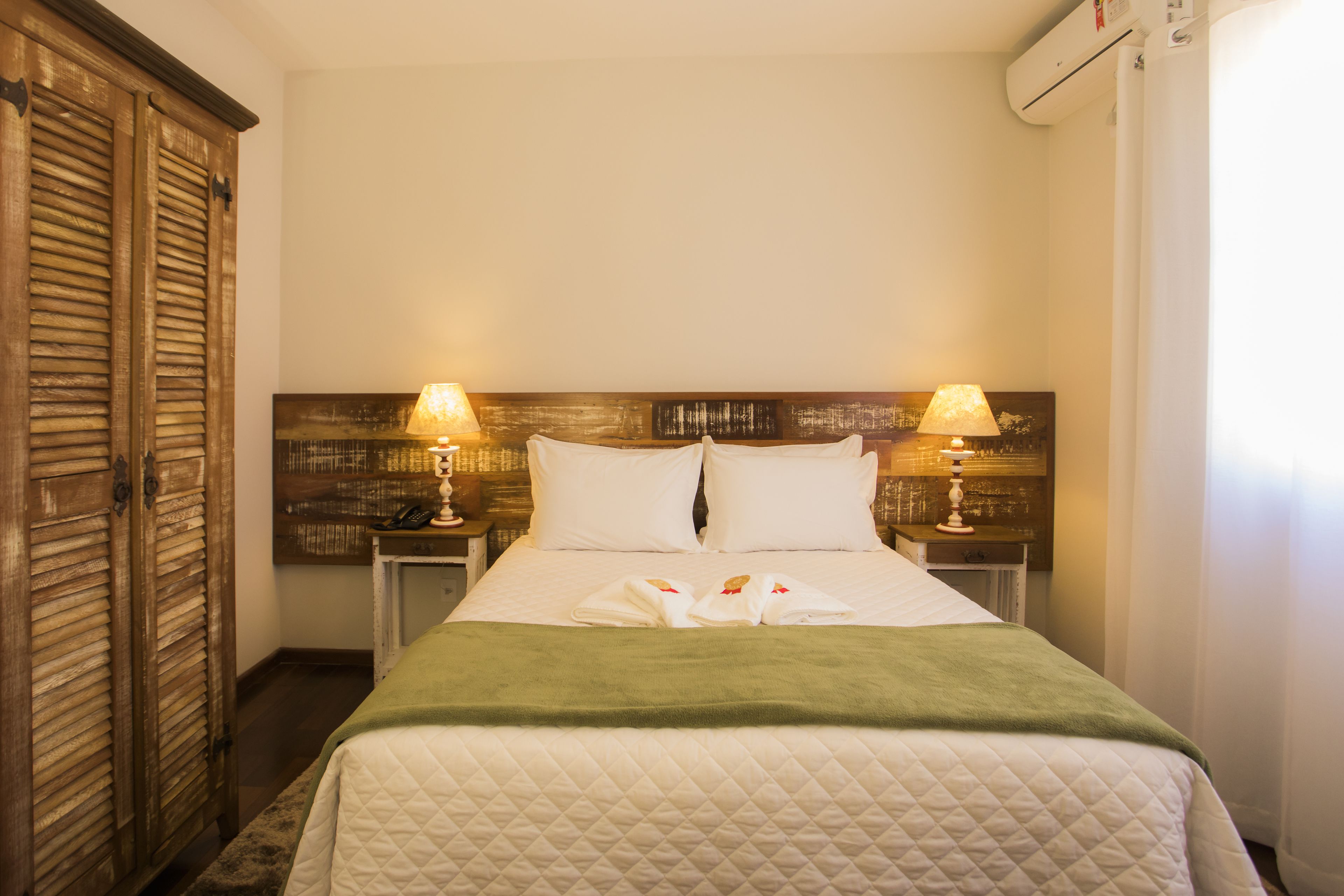 A hotel room with a bed, two bedside lamps, a wooden headboard, and a green throw blanket.