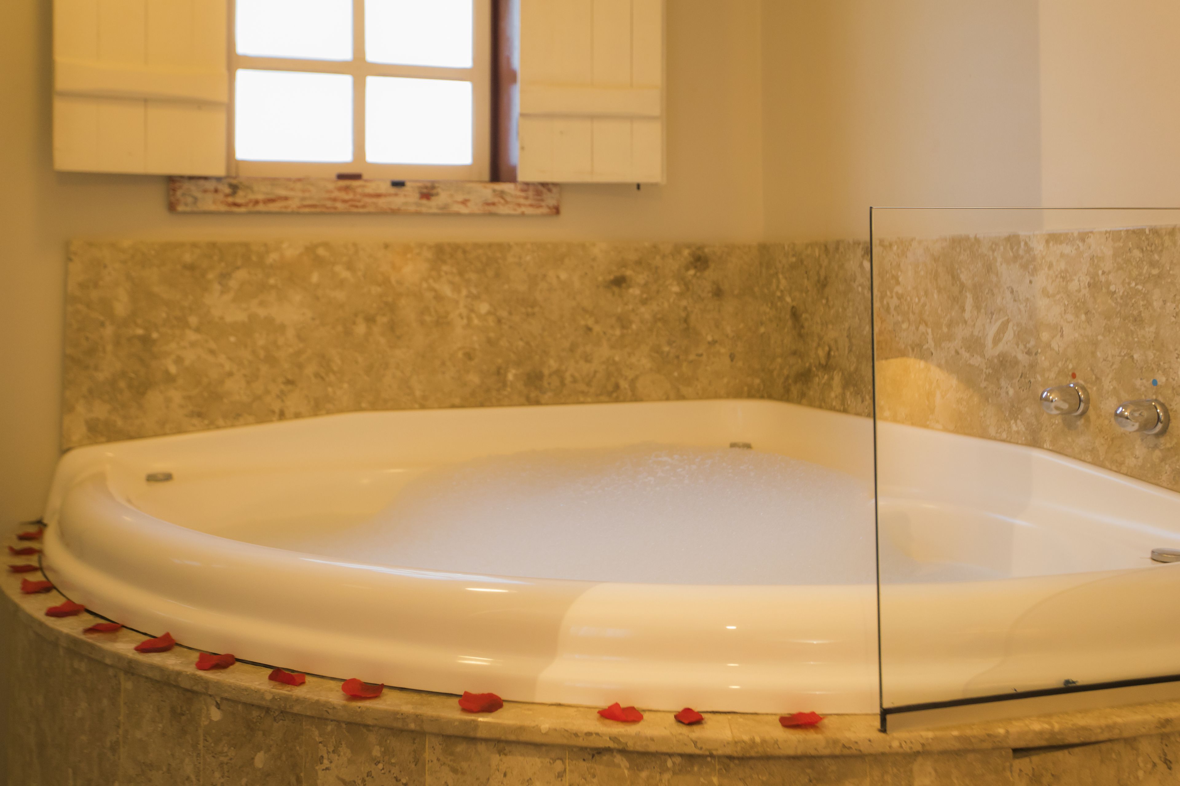 A bathtub with rose petals around the edge, a glass shower enclosure, and a stone wall.