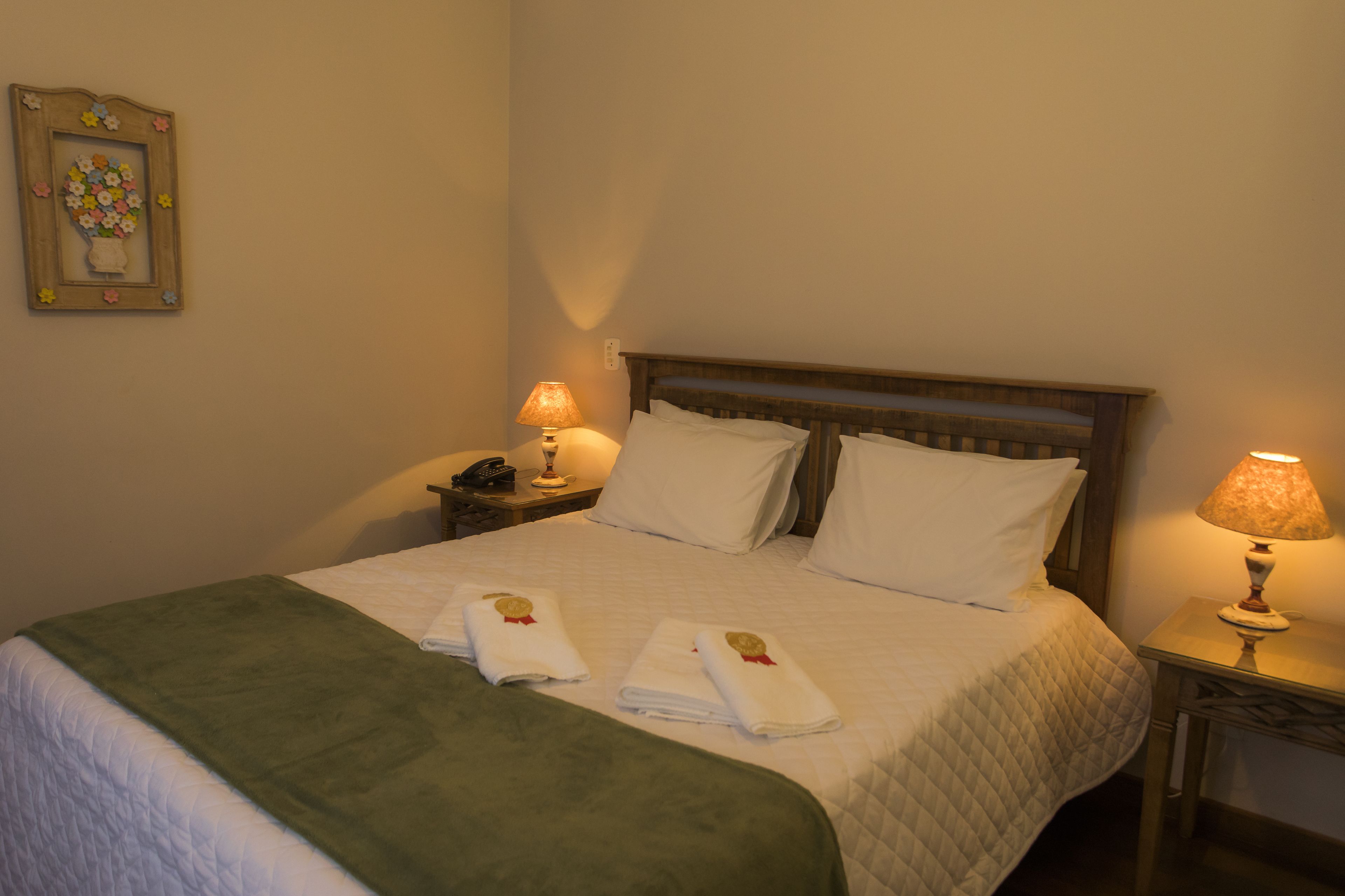 A neatly made bed with white linens, two pillows, and two folded towels with a red ribbon on a green bedspread. There are two bedside tables with lamps, a framed picture on the wall, and a wooden headboard.