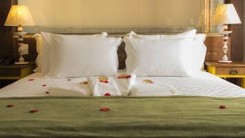 Una cama bien hecha con sábanas blancas, una manta verde y pétalos de rosa esparcidos sobre la cama. Hay dos lámparas de noche sobre mesitas de madera.