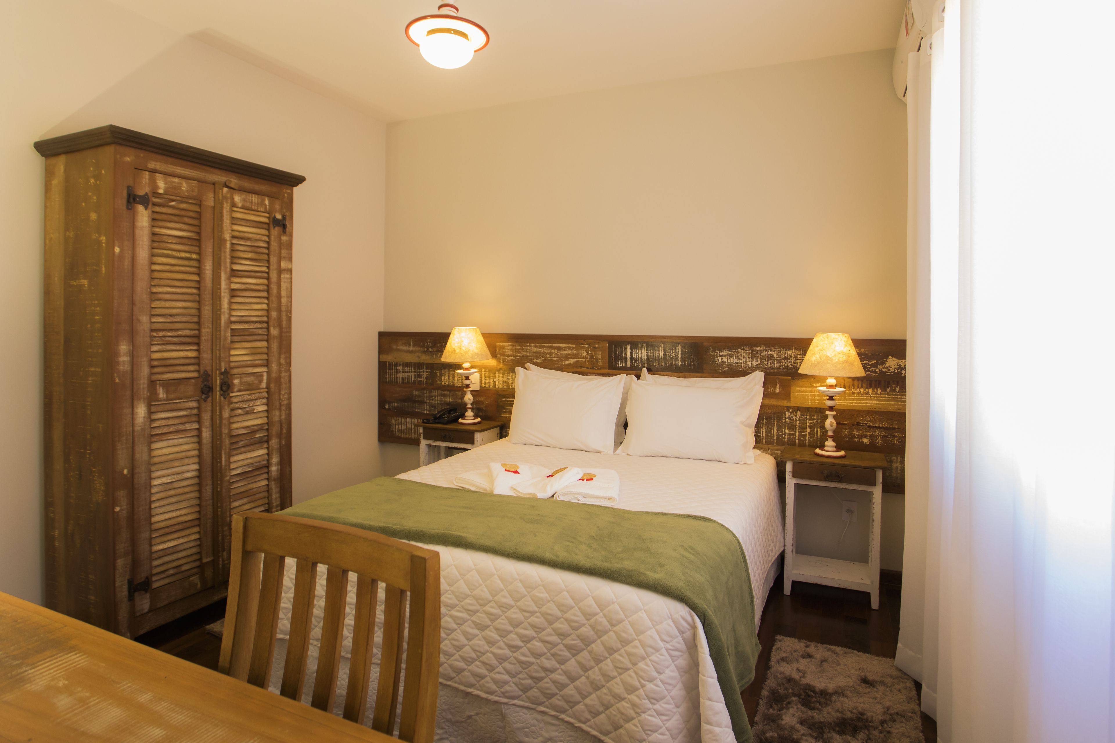 A bedroom with a bed, two bedside lamps, a wooden wardrobe, and a chair.