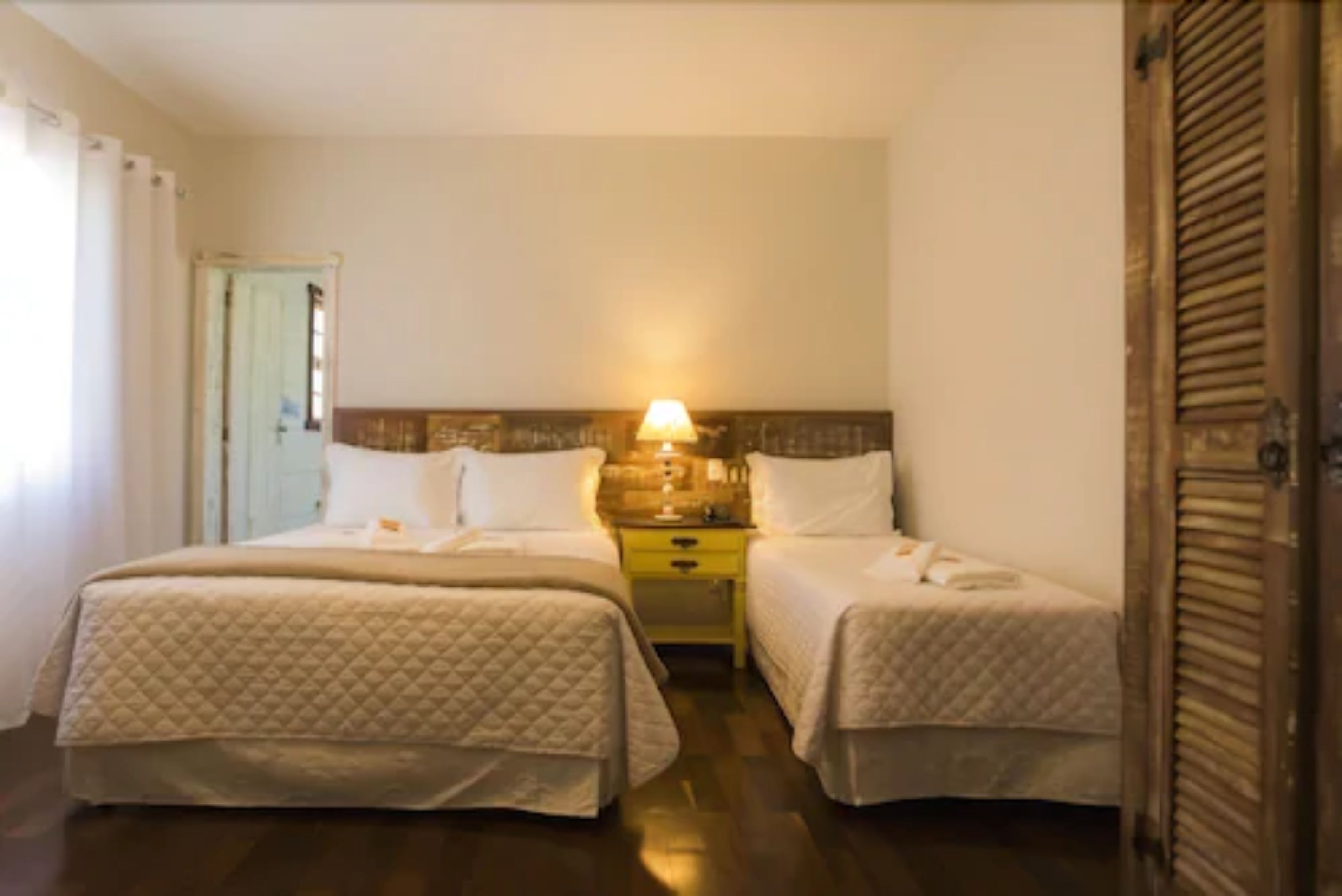 Een hotelkamer met twee bedden, een nachtkastje met een lamp en een deur met gordijnen.
