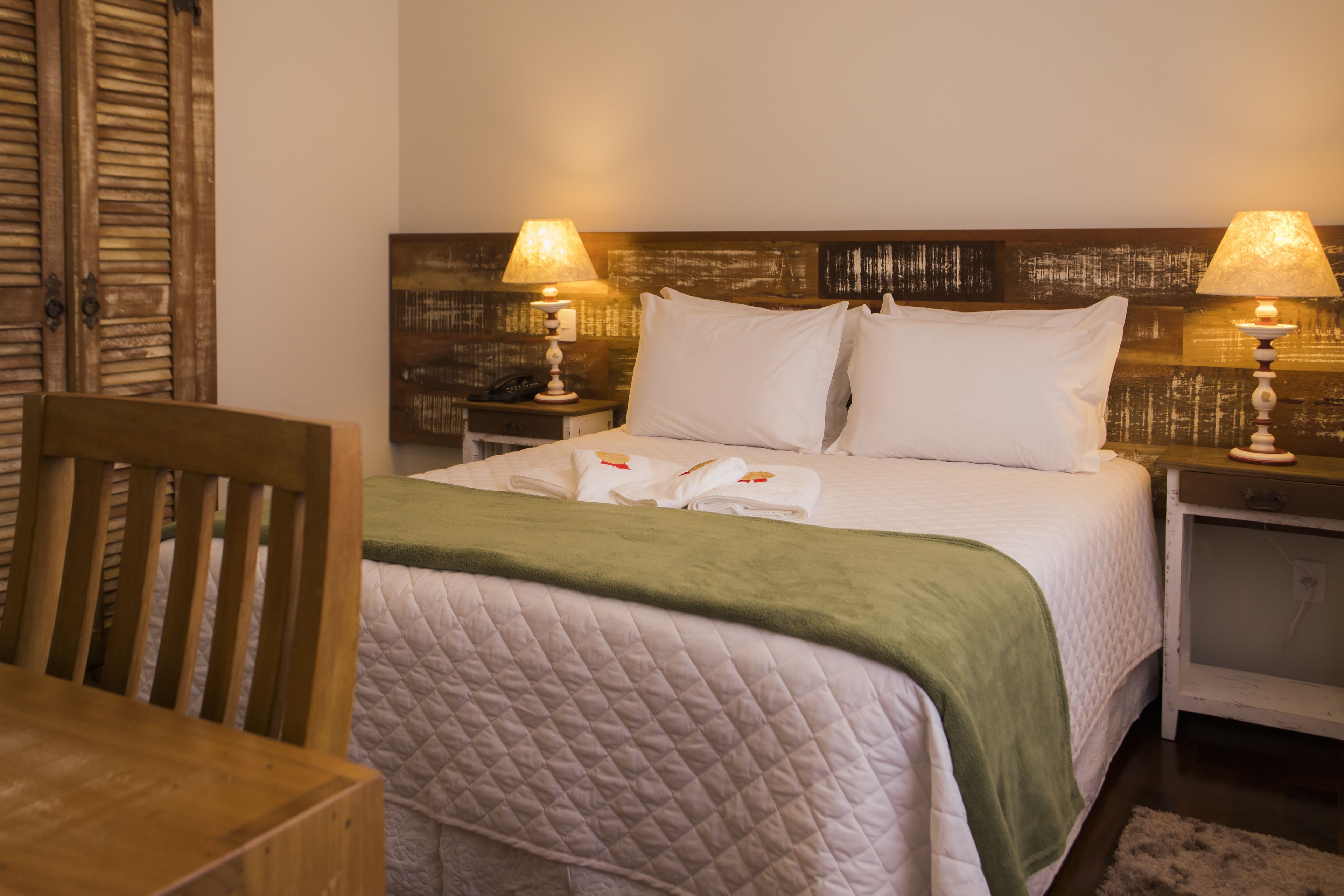 A neatly made bed with a quilted cover, two bedside lamps, a wooden chair, and a nightstand with a lamp.