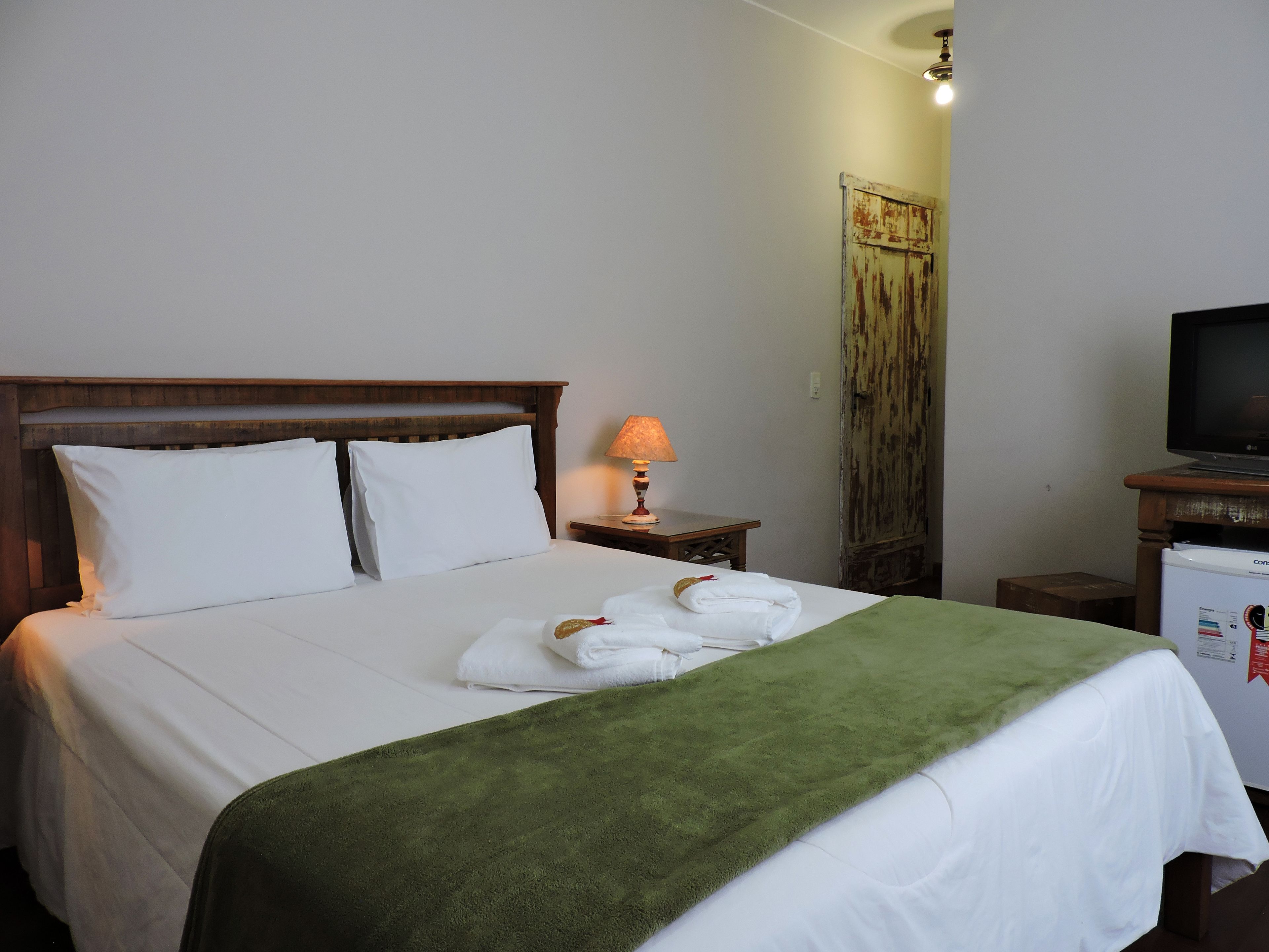 A neatly made bed with white linens and a green blanket, a bedside lamp, and a television on a wooden stand.