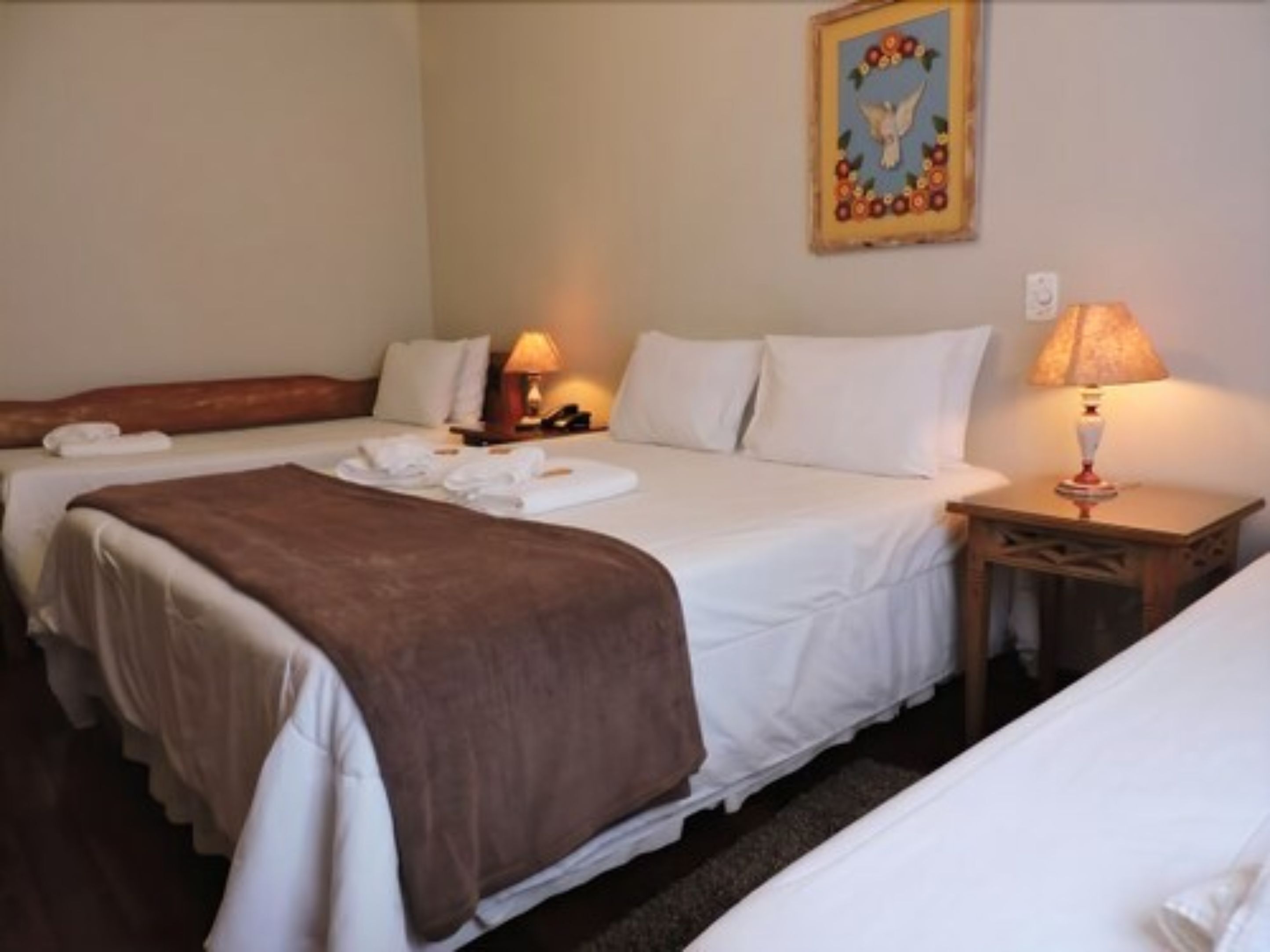 A hotel room with two beds, white linens, a brown throw blanket, a nightstand with a lamp, and a painting on the wall.