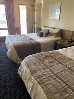 Una habitación de hotel con dos camas, cabecero de madera, mesita de noche con un reloj y una ventana con cortinas.