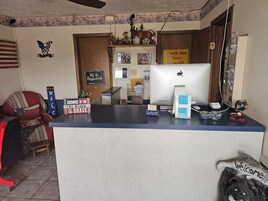 Un mostrador de recepción con una computadora, un letrero con la contraseña de 'Wi-Fi' y diversas decoraciones, incluyendo un cartel que dice 'Hogar de los valientes' y otro que dice 'Bienvenido'.