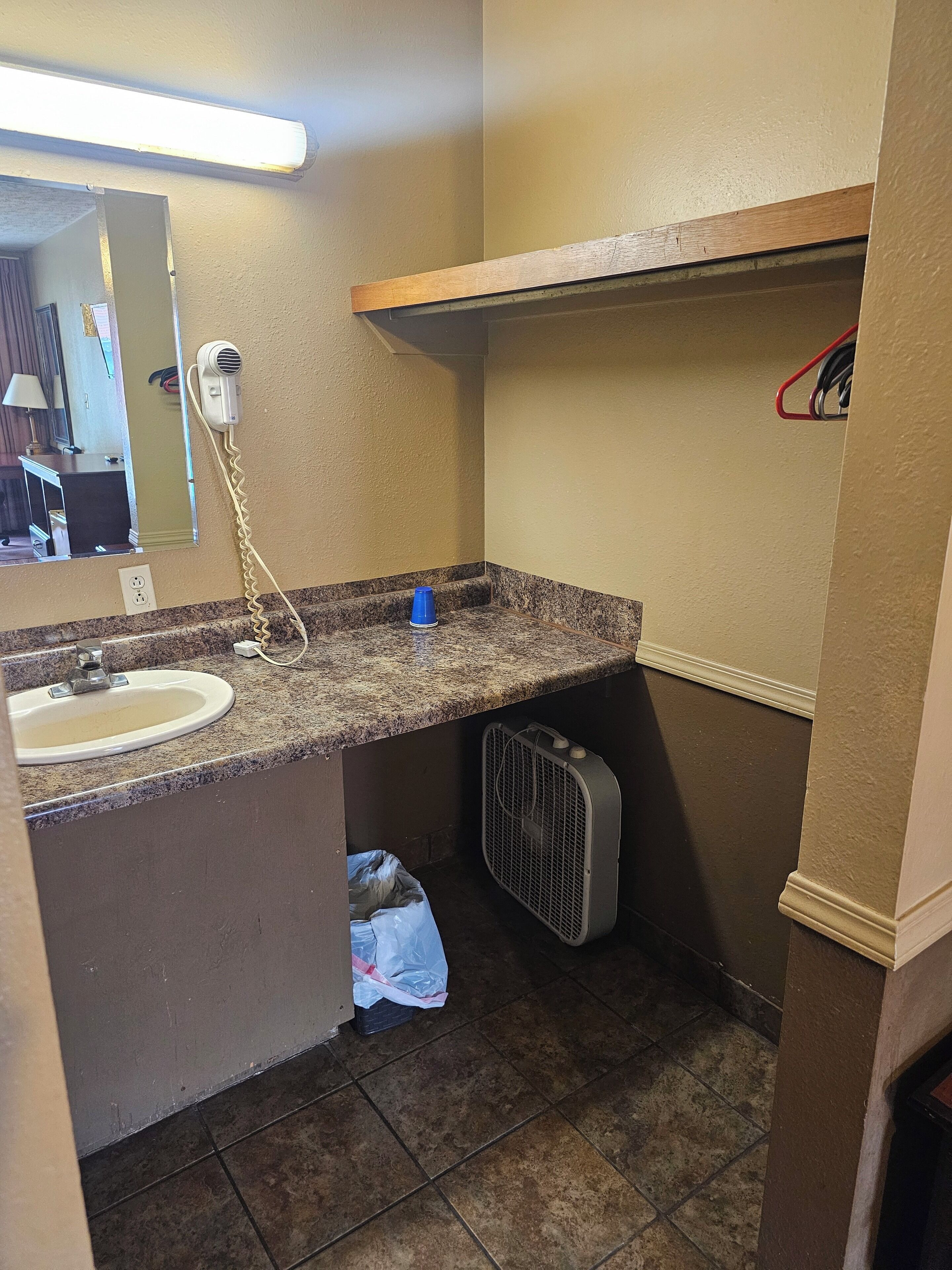 A bathroom with a sink, mirror, and a hairdryer plugged in. There is a blue cup on the counter and a trash bin under the sink.