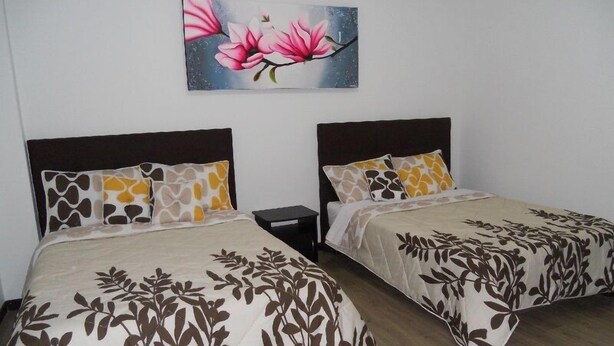 Two single beds with dark headboards and patterned bedspreads, a nightstand, and a painting of pink flowers on the wall. - Apartamento Quinta Paredes 036 (Bogotá)