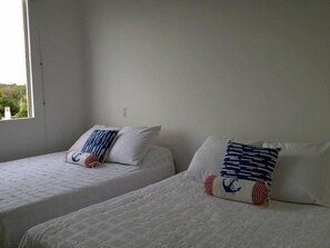 Two single beds with white bedding and patterned pillows, a window with a view of greenery, and a plain white wall.