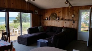 A cozy room with a wooden interior, a large window offering a view of the sea, a dark-colored sofa, and a small table with chairs. -  LAKE MICHIGAN BEACHFRONT! PRIVATE BEACHFRONT! DISCOUNTED SEASONAL RATES  (Montague)