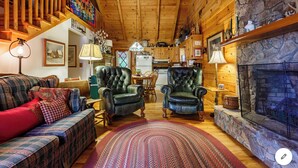 A cozy log cabin interior with a fireplace, two leather armchairs, a plaid sofa, and a dining area.