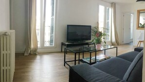 Uma sala de estar moderna com uma televisão de tela plana sobre uma mesa de centro de vidro, um sofá cinza e um vaso com planta.