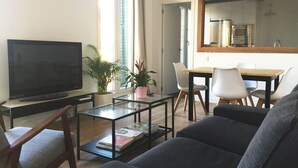 Uma sala de estar com televisão de tela plana, uma mesa de centro de vidro, um sofá e um espaço de jantar com cadeiras e mesa.