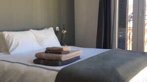 Uma cama bem arrumada com lençóis brancos, um cobertor cinza e uma pilha de toalhas dobradas.