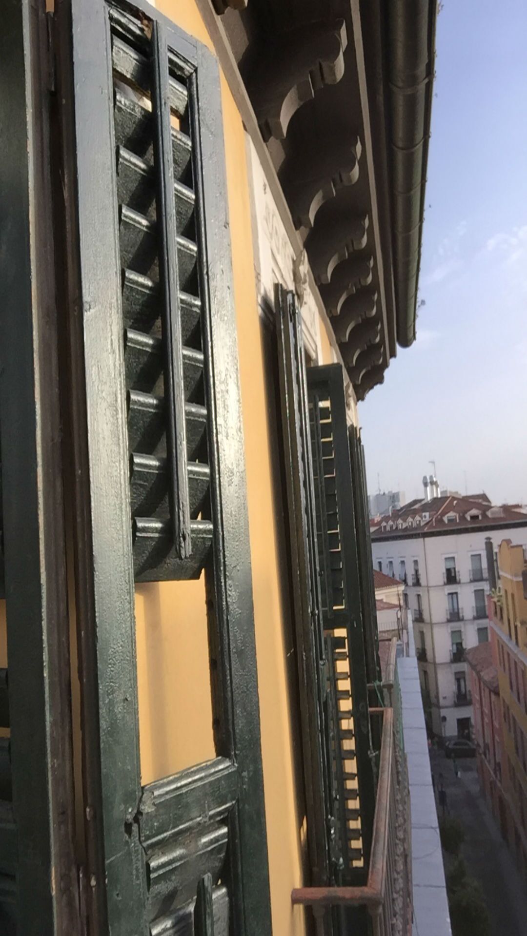 A balcony with black shutters and a yellow wall, overlooking a cityscape with buildings.
