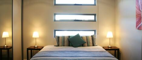 A hotel room with a large bed, two bedside tables with lamps, and three windows.