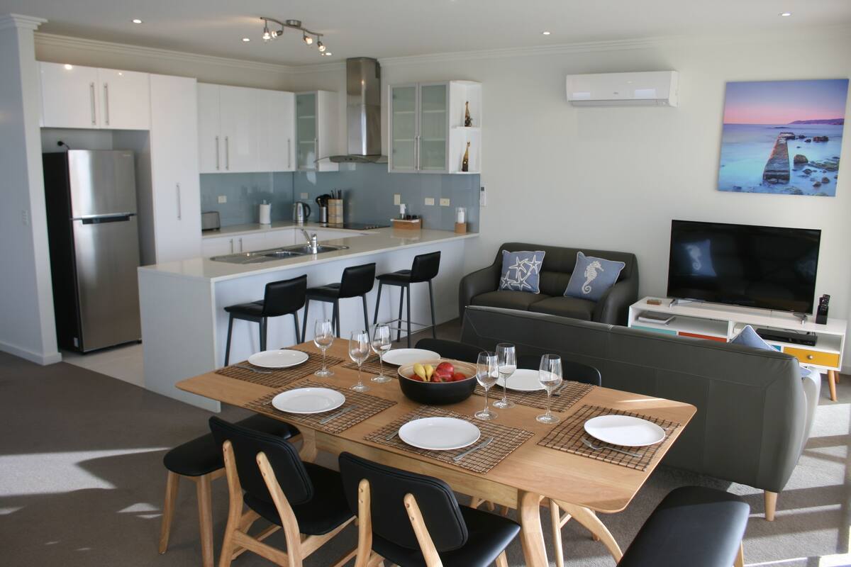 Una cocina moderna con armarios blancos, un refrigerador de acero inoxidable y una zona de comedor con una mesa de madera dispuesta para cuatro.