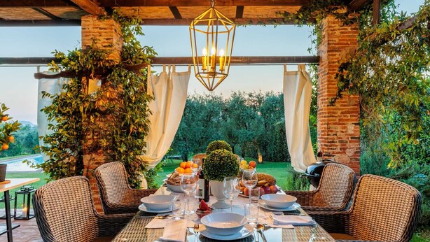 Un espace extérieur aménagé pour les repas, avec des meubles en osier, une table dressée et une vue sur une piscine et la verdure.