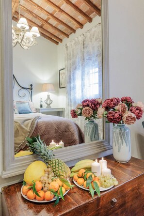 A bedroom with a bed, a nightstand, a mirror, and a dresser. There are flowers on the dresser and a fruit bowl on the dresser.