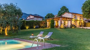 Un jardin bien entretenu avec une piscine, deux chaises longues et une pergola agrémentée d’un éclairage extérieur.