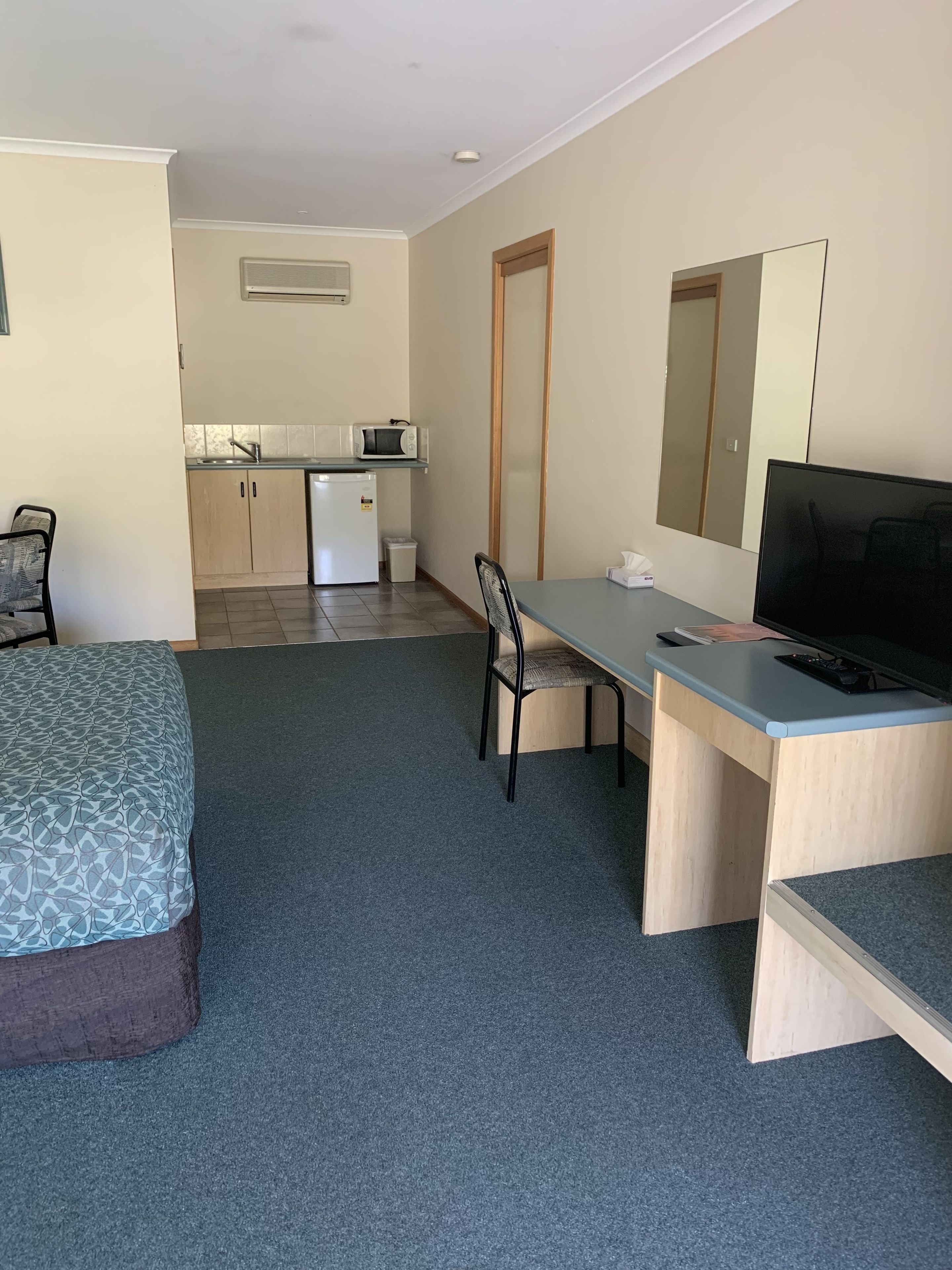 A hotel room with a bed, a desk with a chair, a television, a small kitchenette, and a mirror.
