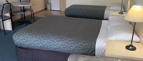 A hotel room with a large bed, a small kitchenette, a dining table with chairs, and a single bedside lamp.