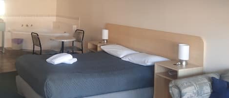 Habitación de hotel con cama, mesitas de noche, una silla, una pequeña mesa y un baño al fondo.