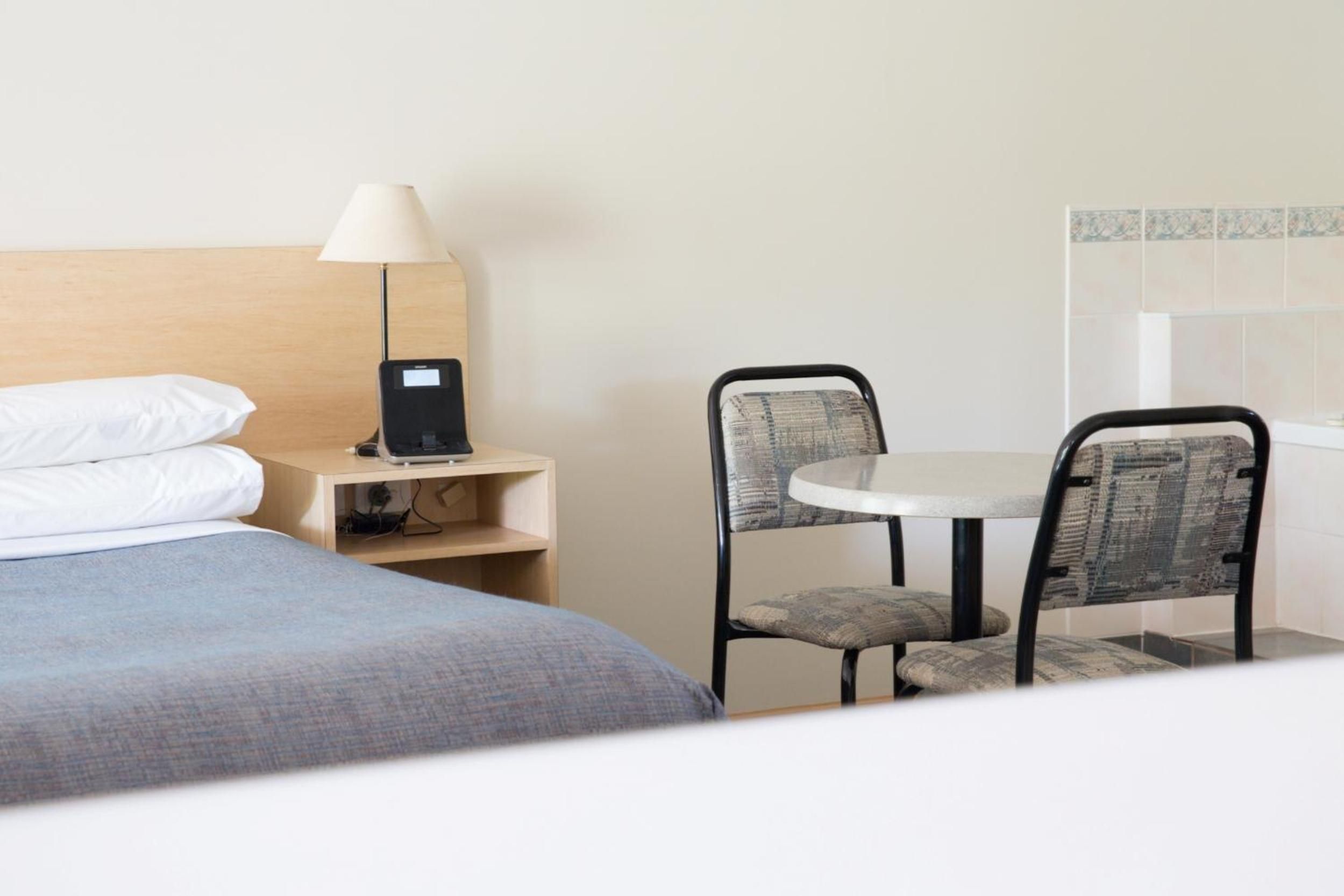 A hotel room with a bed, a desk with a lamp, a small table with two chairs, and a wall-mounted control panel.