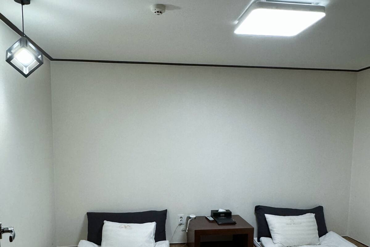 Une petite chambre propre, avec deux lits, une table de chevet, un bureau et un luminaire au plafond.