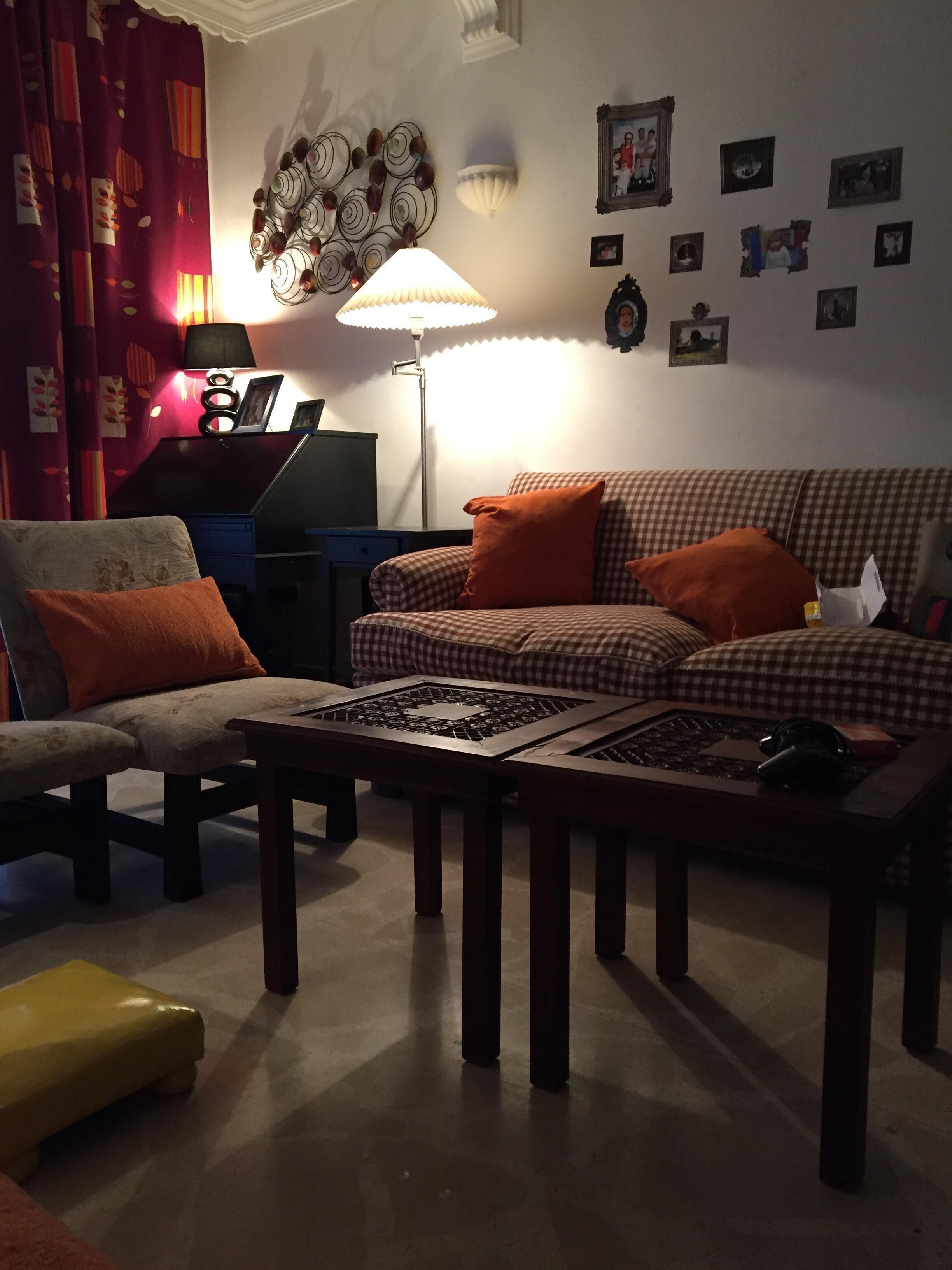 A living room with a sofa, armchair, and coffee table. There is a lamp, a laptop on a desk, and various wall decorations.