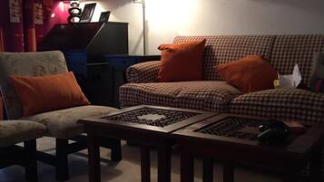 A living room with a sofa, armchair, and coffee table. There is a lamp, a laptop on a desk, and various wall decorations.