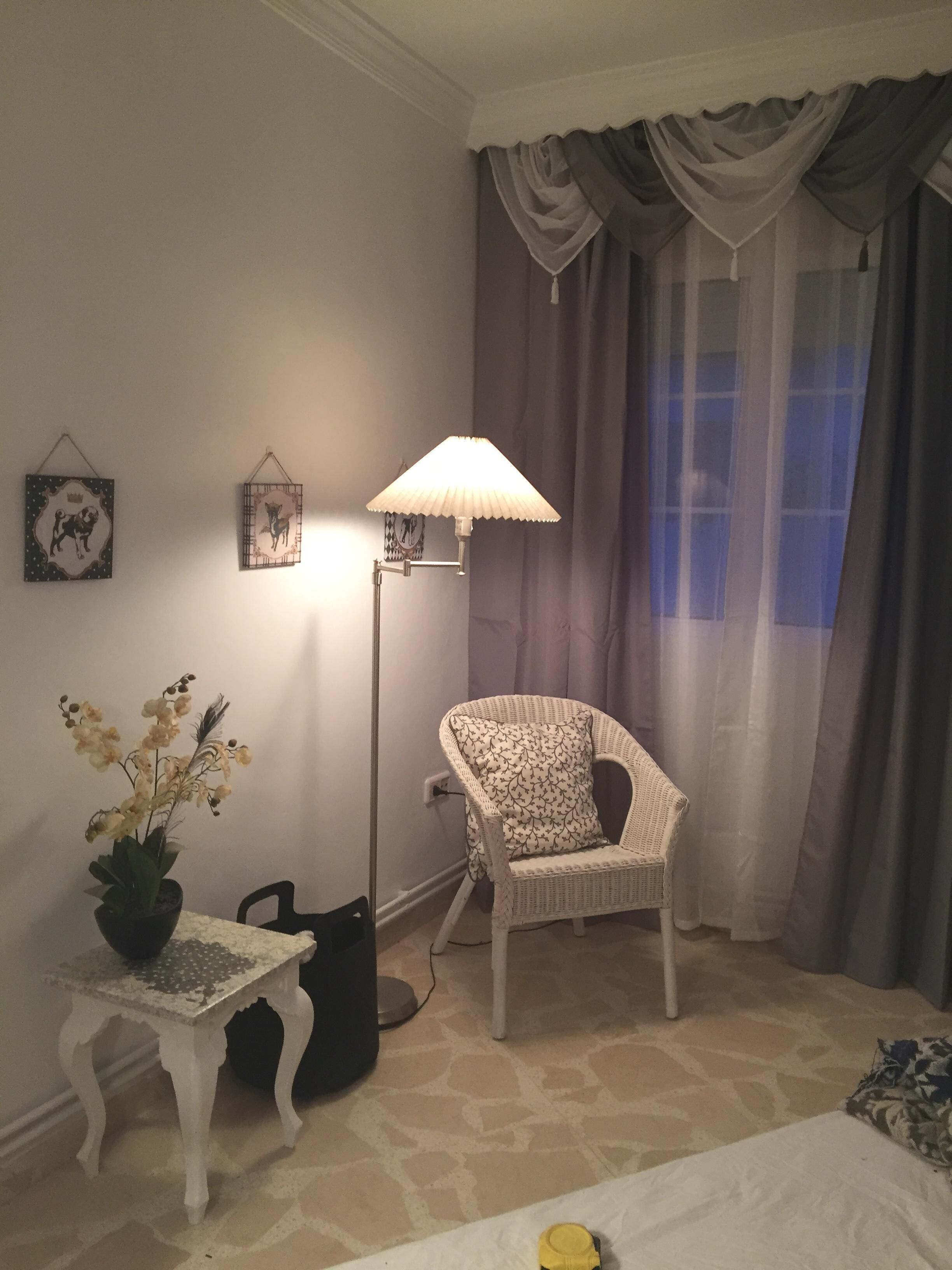 Una habitació amb una cadira de vímet blanc, una taula petita amb una planta en testos, una làmpada de peu i un llit amb un coixí groc.