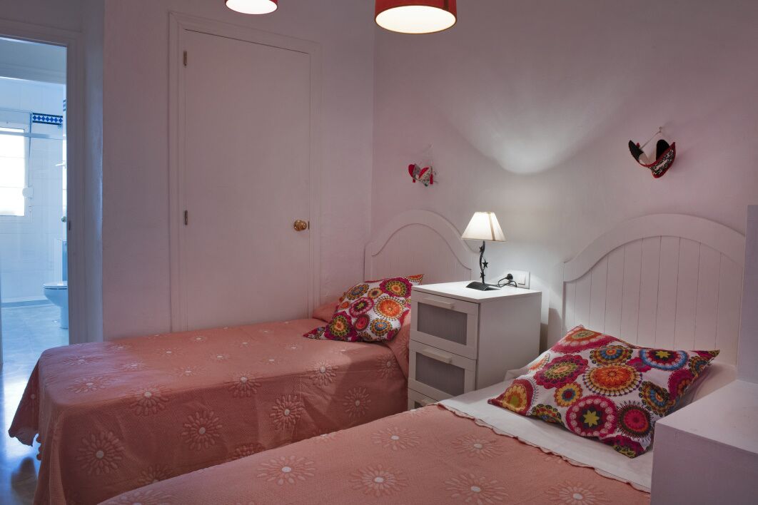 Quarto com duas camas, cada uma com colcha rosa e almofadas coloridas. Há uma mesa de cabeceira com abajur e uma pequena prateleira. O quarto tem paredes brancas e uma porta à esquerda.