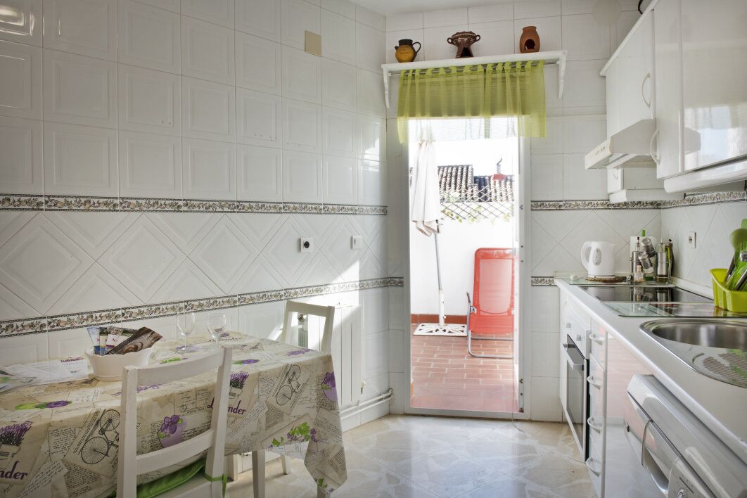 Une cuisine avec des meubles blancs, une table à manger recouverte d’une nappe à motifs floraux et une chaise rouge.