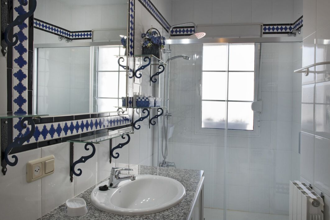 Un baño con un lavamanos blanco, un espejo con detalles de azulejos azules, una ducha y una ventana.