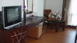 A hotel room with a wooden desk, a television, a chair, a small table with flowers, and a large mirror. - Grand Southern Hotel (Thung Song)