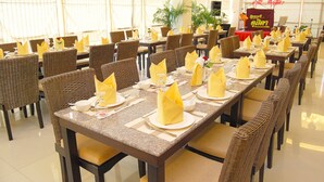 A dining area with long tables set with yellow napkins, plates, and cutlery. There are wicker chairs and potted plants in the background. - Grand Southern Hotel (Thung Song)