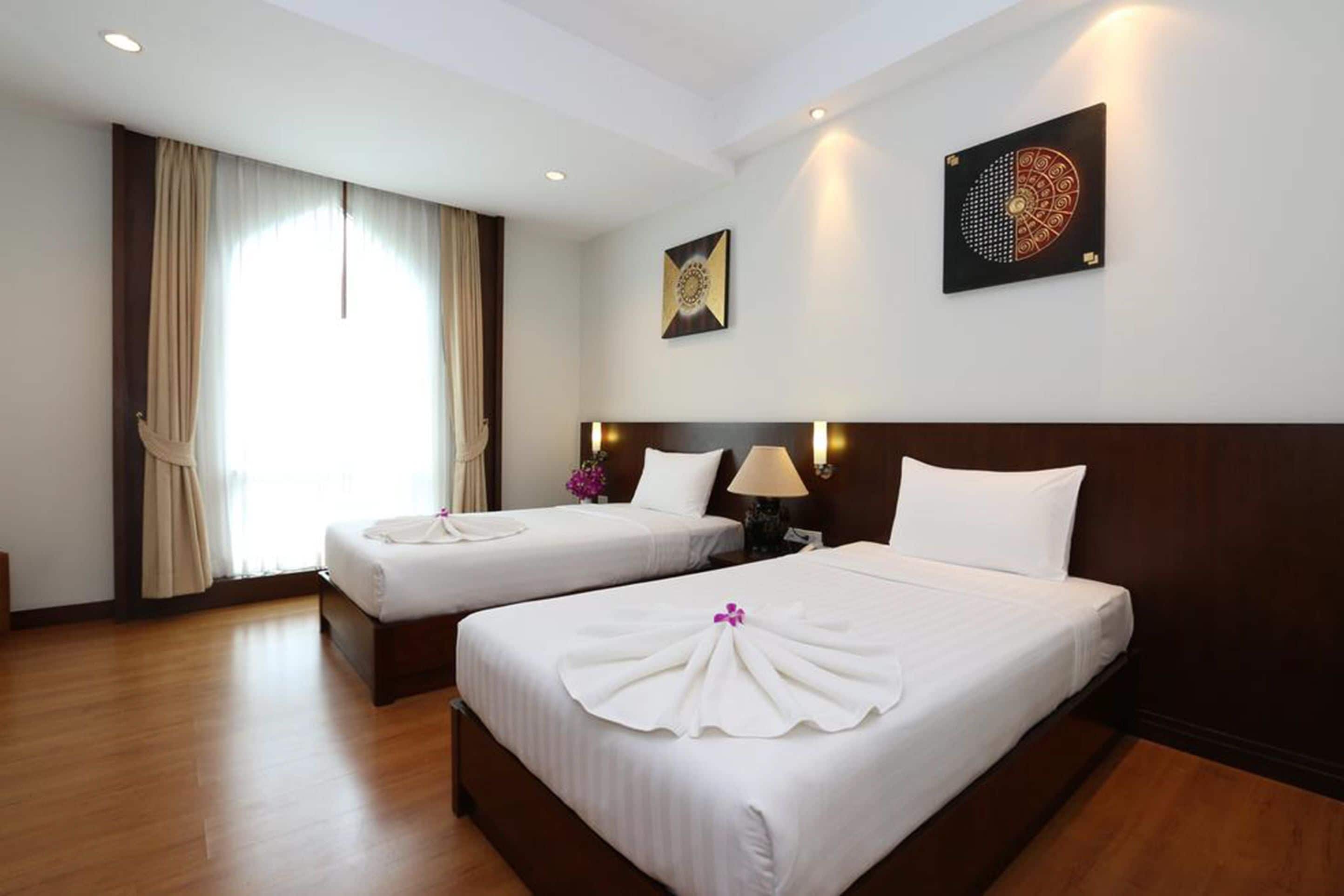 A hotel room with two beds, a large window with curtains, a wall-mounted artwork, and a nightstand with a lamp.